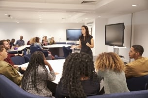 A female instructor stands in front of a small class, explaining a concept.