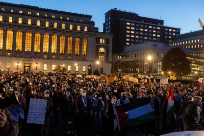 A nighttime scene of a large mass of students attending a pro-Palestinian rally on Columbia University's campus, with Butler Library lit up in the background.