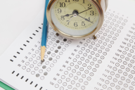 A standardized test answer sheet with bubbles filled in. A pencil and a small circular clock sit atop the sheet.