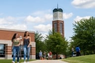Two girls walk on a sidewalk with a college campus behind them. 