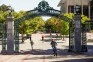 Students riding bicycles and walking near an arched entrance on the UC Berkeley campus