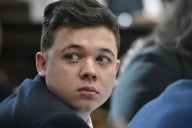 A close up of Kyle Rittenhouse, a young white man with brown hair, glancing behind himself while in a courtroom.