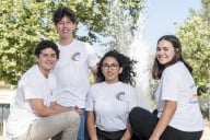Four students pose in white T-shirts, facing the camera, in front of a fountain at Pepperdine University.