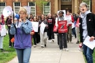 A photograph of Rutgers University demonstrators, including one holding a megaphone and another holding a sign saying “Our Working Conditions Are Students’ Learning Conditions” around a fist grasping a pencil.