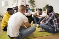 A group of six college students sits on the floor in a living room/common area having what appears to be an engaged, supportive discussion.