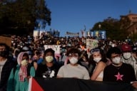 Pro-Palestinian protestors wearing masks and holding signs stand on stairs near an encampment at the UCLA campus 
