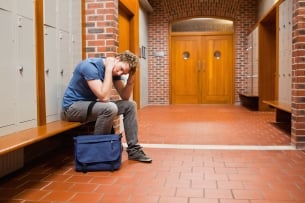 A male college student sits alone in a hallway with his hands on his head, looking anxious and stressed.
