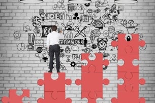 Rear view of businessman standing on red puzzle pieces and drawing business graffiti on brick wall, with the words Teamwork and Idea visible.
