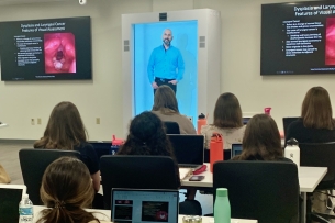 A virtual man appears on a screen in a person-size white box in front of a classroom of students