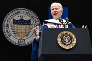 President Joe Biden stands in a blue graduation robe at a podium with the crest for Howard University in the background. 