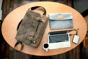 A photo taken from overhead of a shoulder bag, a laptop sleeve, a laptop, an external hard drive and a cup filled with coffee, all atop an oval-shaped table.
