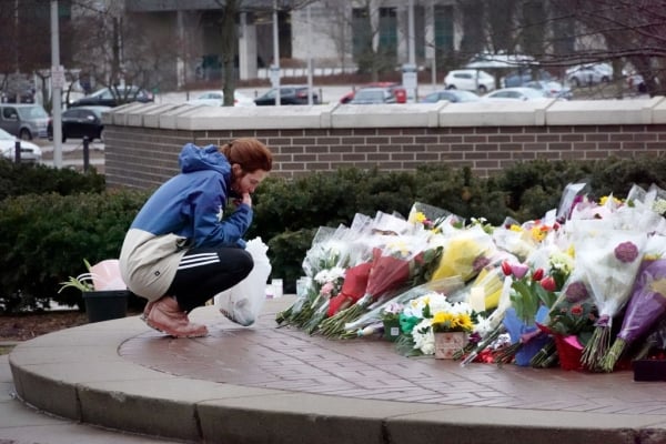 A person in a hoodie crouches next to bouquets of flowers at a makeshift memorial.