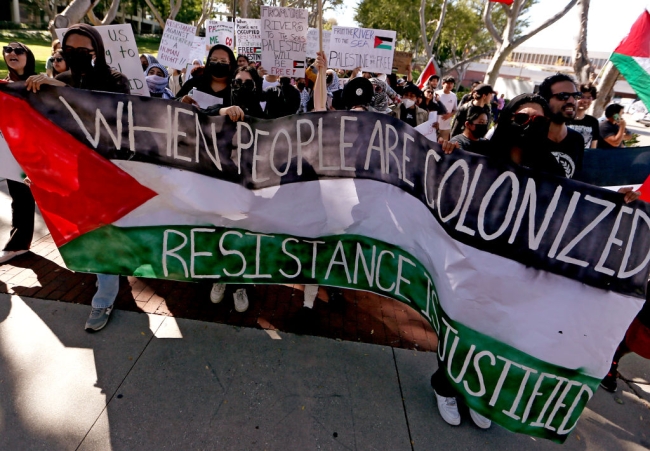 Student protesters have unfurled a large banner that features the Palestinian flag's colors and reads “When people are colonized, resistance is justified.” More protesters can be seen in the background, some holding signs that say things like “from the river to the sea, Palestine will be free” and “resistance against colonization is a human right.”