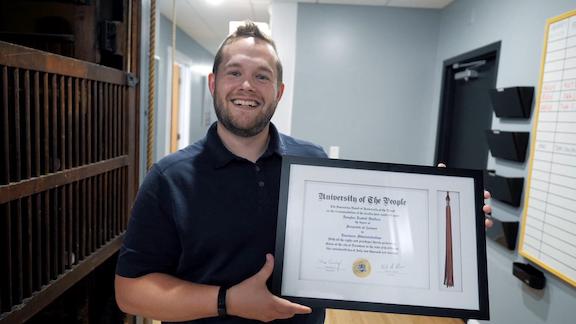 A young man smiling, holding a framed university degree.