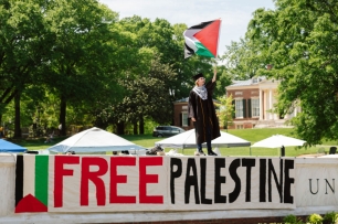 A student in a graduation gown stands above a sign that says “free Palestine.”