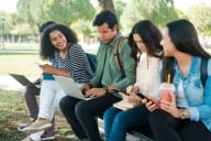 Five students sit outside on a college campus on their laptops and phones.