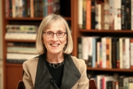 A photo of Claudia Goldin, a light-skinned woman with shoulder-length light hair, pictured in front of a bookcase.