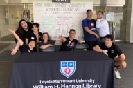 Several students and several more library staff members sitting at a table outside the library at Loyola Marymount University in California to welcome students to the Long Night Against Procrastination event. A whiteboard behind them details the event. Most people in the photo are in humorous poses or making funny faces.