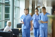 Three students in blue scrubs walk down a hallway in a hospital