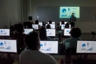 A group of students sits in front of computers, each showing charts and graphs on the screen. They are all facing toward the front of the room with their backs turned toward the camera.
