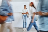 A lone female college student stands in the background of the photo. In the foreground, three students, their photos blurred, suggesting they have places to go, walk past her.