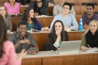 A multi-ethnic group of college-aged students sit in a row in a lecture hall and listening to and apparently enjoying their professor whose back is turned to the camera.