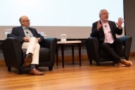 Two men sit on a stage at a conference. The man on the right is speaking and gesturing with his hands.