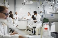 A group of students and a professor meet in a research laboratory. All are wearing lab coats.