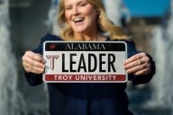 A blonde woman holds a customized Alabama license plate for Troy University with the vanity tag “leader” in all caps
