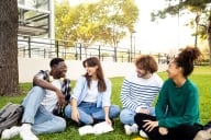 Group of diverse college student friends laughing and chatting sitting on the grass on campus