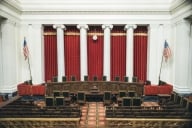 A photo of the interior of the U.S. Supreme Court chambers, showing a curved table with nine chairs in front of a set of five long red curtains hanging between four white columns.
