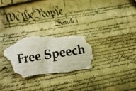 A cutout of a piece of paper with the words "Free Speech" laid atop a sepia-toned copy of the U.S. Constitution.