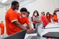 Two incarcerated students wearing masks look at a laptop with classmates talking behind them. 
