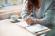 woman in cozy sweater sits before a window writing in a notebook with a coffee cup by her side