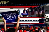 Former president Trump stands in front of a plane at a podium. A Trump 2024 sign is in the foreground.
