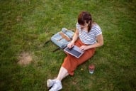 Woman sits on grass typing into a computer with a satchel and a paper cup of coffee on either side