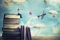 Woman sits atop a high stack of old books looking through a telescope at bright colored flying balloons and an airplane