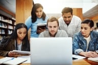 Five students gather around a laptop in a university library.