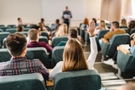 Back view of female college student raising her hand to answer the question in a class lecture hall.