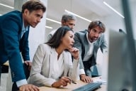 Asian woman sits at a desk looking at a computer with concern, while three men stand behind her also looking worried