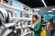 A young woman wearing a yellow backpack reads a book in a campus library