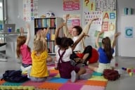 A teacher sits on the floor with a group of young students, all with their hands in the air. 