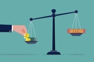 Hand placing money on one side of a scale weighing down the word "justice" on the other side of the scale