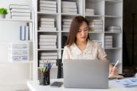Asian woman sits in front of a bookcase and at a desk with a computer and charts 