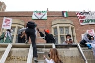 Pro-Palestinian protestors trying to set up a solidarity encampment at the University of California, Los Angeles's Kerckhoff Hall May 23. One protester climbs a ladder to what appears to be a roof to join other protesters; signs in the background espouse support for Palestine.