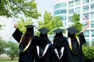 Rear view of four female graduates wearing graduation caps, gowns and hoods on campus.