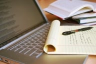 A notepad, the first page filled with handwritten notes, lies atop an open laptop, next to a stack of books.