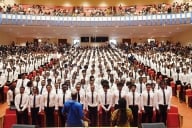 Students, in white shirts and dark neckties, crowd an auditorium