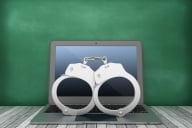 Computer with handcuffs on top of it sits in front of a blackboard 