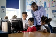 A low-angle view of a teacher and his students learning in a classroom. A little boy in red is sitting at a desk showing the group his work on the computer.